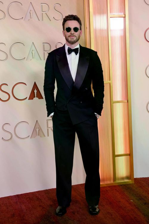 Chris Evans xuất hiện trong bộ tuxedo đen hai hàng khuy của Giorgio Armani. Nam diễn viên kết hợp kính râm tròn và nơ đen, tạo phong cách vừa cổ điển và mang lại vẻ lịch lãm, đậm chất quý ông Hollywood.