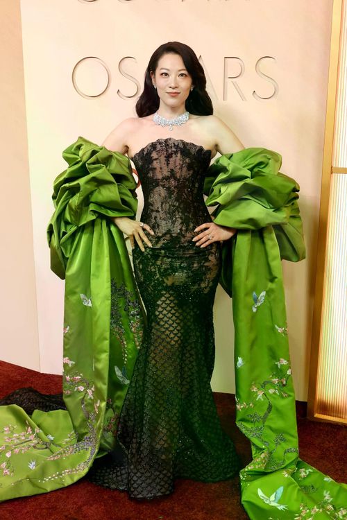 Arden Cho xuất hiện trong thiết kế của Miss Sohee Couture. Bộ váy ren đen ôm sát kết hợp áo choàng màu xanh lá thêu họa tiết hoa và chim, tạo nên vẻ ngoài sang trọng và nổi bật trên thảm đỏ.