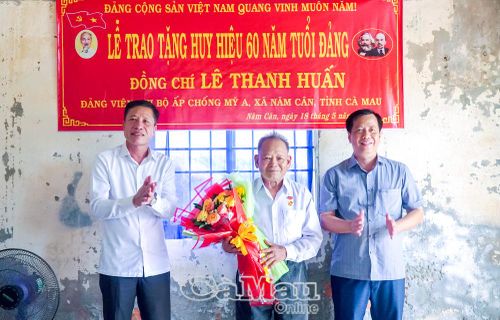 Đồng chí Trần Hữu Phước, Trưởng ban Nội chính Tỉnh ủy và đồng chí Huỳnh Minh Kiên, Bí thư Đảng ủy, Chủ tịch HĐND xã Năm Căn, trao Huy hiệu 60 năm tuổi Đảng và tặng hoa chúc mừng đồng chí Lê Thanh Huấn.