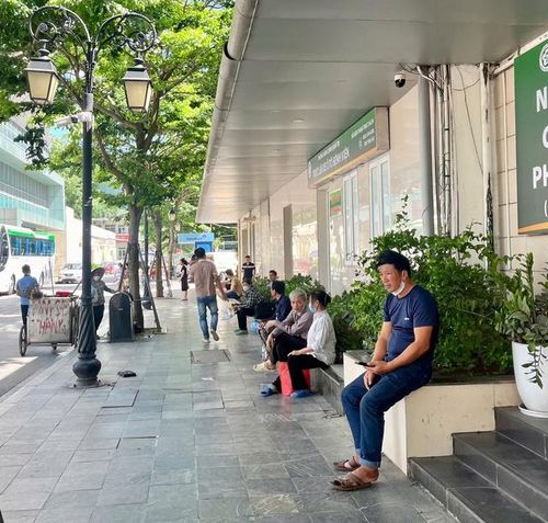 Nhiều người nhà bệnh nhân mang theo đồ đạc, tranh thủ nghỉ ngơi tại các khu vực mát, có gió, ghế đá dưới bóng cây và mái bạt để tránh nắng.