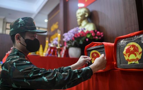  Trong không gian trang nghiêm, một cán bộ Đội Quy tập hài cốt liệt sĩ cẩn thận dán Quốc huy lên tiểu quách chứa hài cốt. Mỗi tiểu quách được bọc vải đỏ, sắp đặt ngay ngắn bên cạnh bàn thờ Chủ tịch Hồ Chí Minh và Quốc kỳ trong Nhà Tưởng niệm. Đây là bước cuối trong quy trình an vị hài cốt trước khi lấy mẫu giám định ADN và lưu giữ theo quy định. 