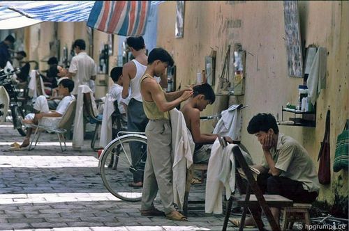  Phố cắt tóc trên vỉa hè phố Quang Trung, đoạn giữa phố Bà Triệu và Trần Quốc Toản, vào khoảng năm 1991-1993. Những ô gạch lát vỉa hè, bức tường vàng cũ kỹ cùng các quầy cắt tóc đơn sơ tạo nên khung cảnh bình dị, đặc trưng của Hà Nội thời bấy giờ. Mỗi quầy chỉ có một chiếc gương treo tường, vài chiếc kéo, lược, một ghế gỗ cho khách và đôi khi thêm lọ phấn rôm hoặc keo vuốt tóc tự pha. Khách chủ yếu là nam giới với những kiểu tóc đơn giản. Một lần cắt tóc hoặc cạo râu diễn ra nhanh chóng, chi phí chưa đến một USD. 