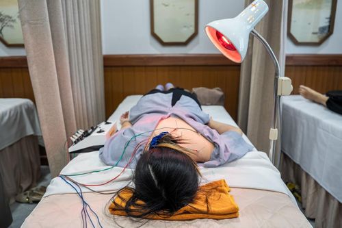  Nằm trên giường điều trị, Hiền được bác sĩ thực hiện điện châm kết hợp chiếu hồng ngoại để giảm đau cổ vai gáy. Điện xung truyền qua các kim châm tác động lên huyệt và các nhóm cơ co cứng, trong khi nhiệt từ đèn hồng ngoại giúp tăng tuần hoàn tại chỗ, hỗ trợ thư giãn cơ và cải thiện khả năng vận động. 