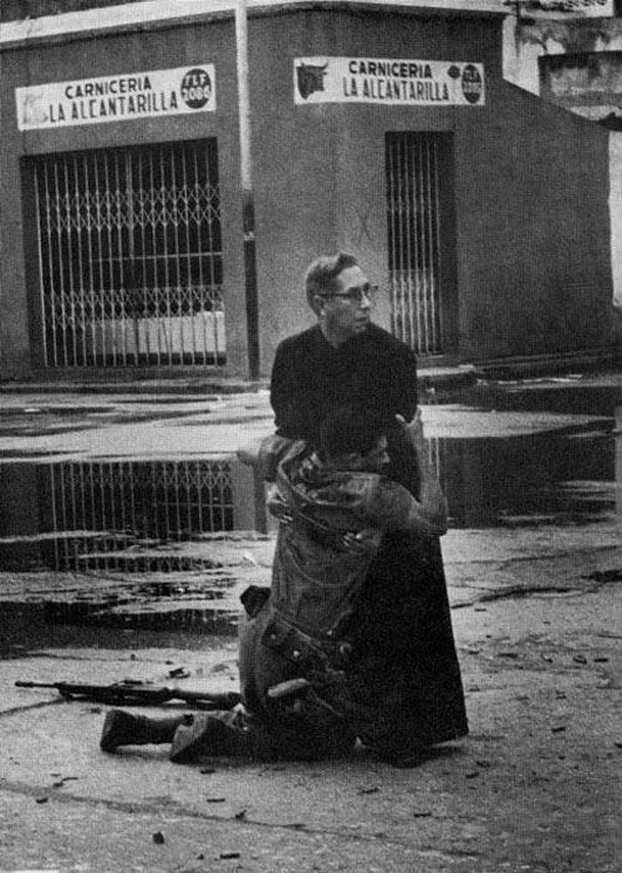 12. Padillo - 1962: Cha tuyên úy phục vụ trong quân đội Venezuela - Luis Paddillo - thực hiện nghi lễ cuối cùng cho một người lính đã bị thương chí mạng. Bức ảnh được chụp ngày 4/6/1962. Cha Padillo ngày hôm đó đã đích thân tìm đến những người lính bị thương sắp qua đời, để thực hiện những nghi lễ tôn giáo cuối cùng cho họ, trong khi đạn nã điên cuồng xung quanh.
