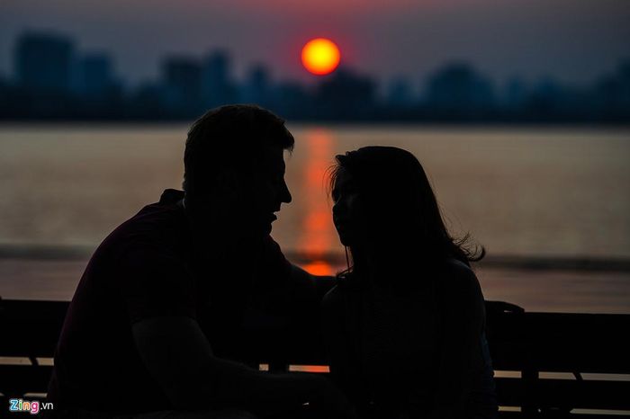 Một chàng trai ngoại quốc ngồi ghế đá ngắm mặt trời lặn cùng cô gái người Việt.