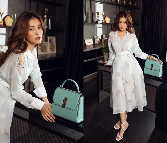 Hoàng Thùy Linh thanh lịch trong thiết kế váy sơ mi thắt eo nữ tính. Tông màu trắng chủ đạo của bộ cánh được điểm xuyết bởi màu vàng nâu của họa tiết thêu nổi bật. Nữ ca sĩ khéo tạo điểm nhấn ánh nhìn với túi xách màu xanh bạc hà cùng tông trang điểm cam sậm vừa tạo sắc thái dịu dàng, vừa mang lại nét lôi cuốn và thu hút cần có.