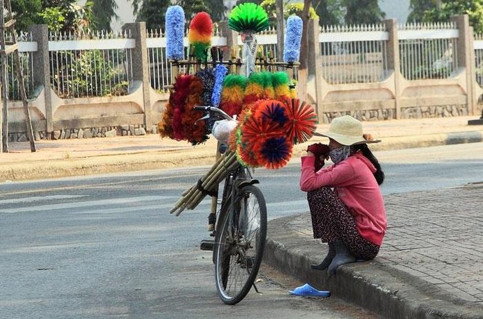  Một người phụ nữ bán các loại chổi dựng xe đạp tránh nắng dưới bóng cây trên vỉa hè. 