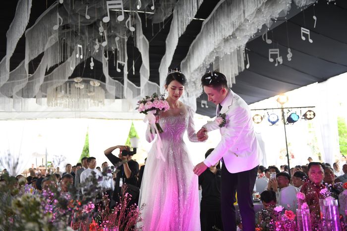  Chú rể Mạnh Quỳnh và cô dâu Khánh Vy tiến vào lễ đường trong tiếng vỗ tay của quan khách. Khoảng 700 khách mời là bà con, hàng xóm tại quê nhà đã đến tham dự và chúc phúc trong ngày trọng đại của cặp đôi. Toàn bộ ghế ngồi của khách đều được trang trí theo tông màu trắng. 