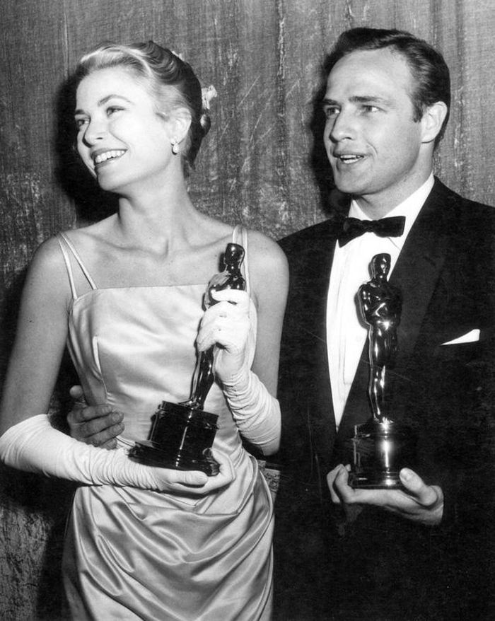 Váy Grace Kelly chất liệu vải satain màu ghi dây đeo vai giản đơn (bên cạnh Marlon Brando) tại gala Oscar 1955.