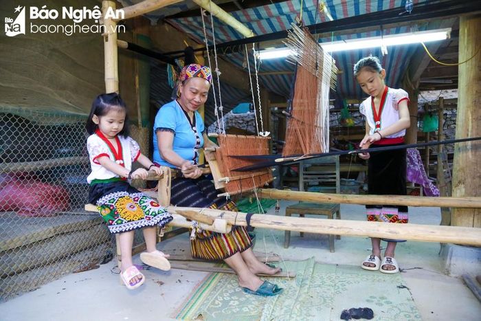  Bà Lô Thị Mai (áo xanh) có 2 cháu gái. Bé lớn là Lô Thị Yến Hương sắp vào lớp 4, bé sau Lô Thị Yến Vi chuẩn bị lên lớp 2. Những tháng Hè vừa qua là quãng thời gian các bé được vui đùa thỏa thích, nhưng bà cũng không quên việc dạy các cháu nghề canh cửi vốn đã trở thành nghề truyền thống của cộng đồng người Thái ở bản Na. Ảnh: Đình Tuyên 