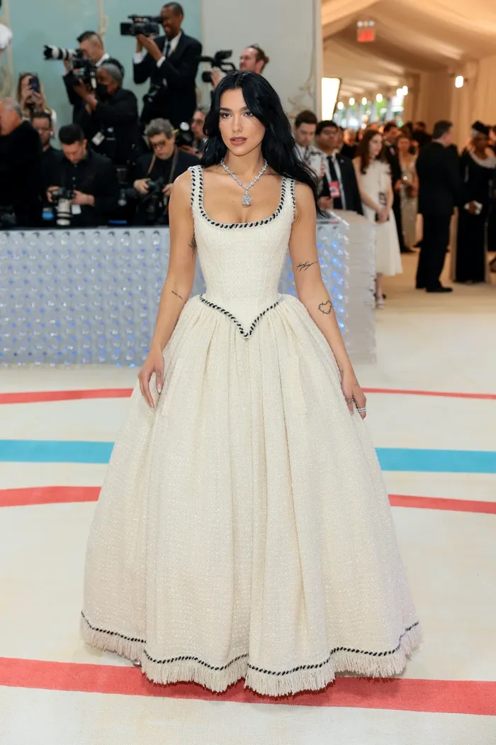Dua Lipa diện bộ đầm vải tweed trắng vintage của Chanel. Thiết kế từng xuất hiện trong show Haute Couture mùa thu năm 1992, do “nàng thơ” Claudia Schiffer thể hiện. Trang phục của cô ghi dấu ấn ở vòng cổ Lucida Star gắn viên kim cương huyền thoại của Tiffany nặng hơn 100 carat, được Vogue mô tả “đẹp đến kinh ngạc”.