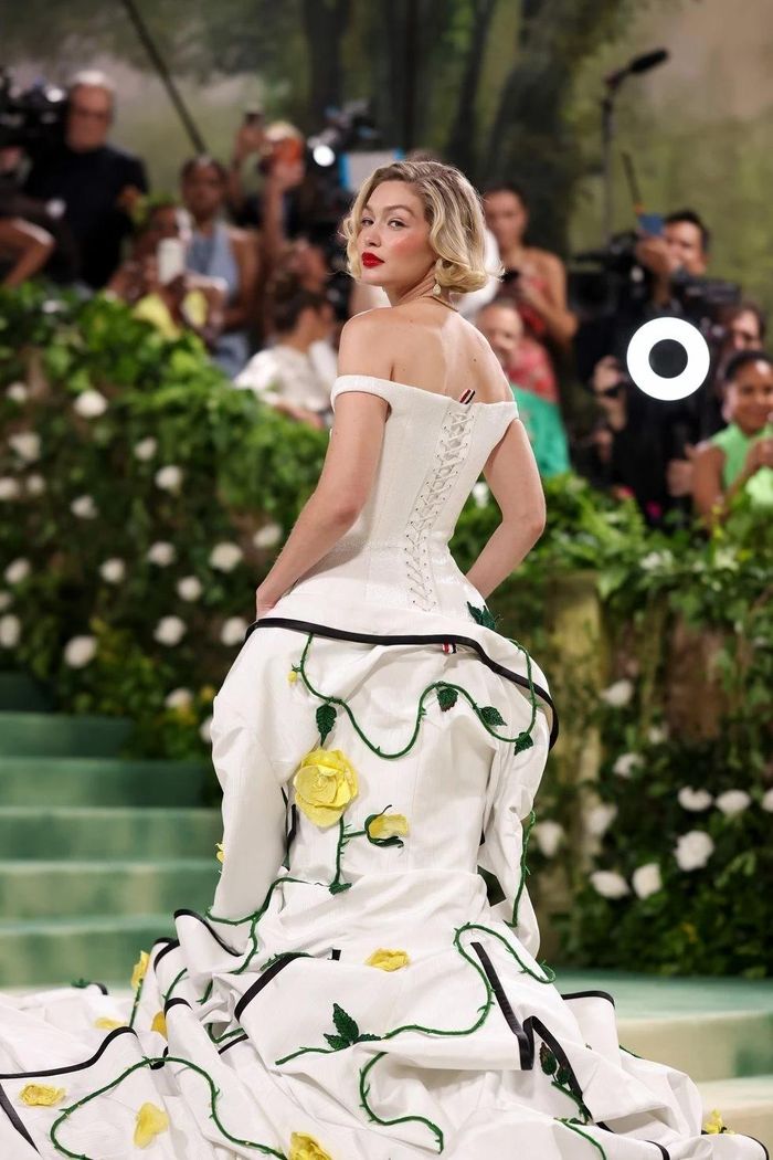  Xuất hiện trên thảm đỏ sự kiện, siêu mẫu Gigi Hadid đem đến tạo hình cổ điển, vừa sang trọng vừa quyến rũ. Cô thực hiện kiểu tóc ngắn vàng hoe, tô son đỏ đậm, gợi nhớ đến biểu tượng thời trang Marilyn Monroe. Nữ minh tinh diện chiếc váy trắng trễ vai của nhà mốt Thom Browne. Điểm nhấn của thiết kế này là những bông hồng vàng 3D được sắp xếp khéo léo trên chân váy. Khu vườn hoa hồng trên mẫu đầm được một đội ngũ gồm 40 người thực hiện trong 8.500 giờ. Sau khi được Gigi mặc trên thảm đỏ, trang phục sẽ được đưa vào kho lưu trữ của Thom Browne. 