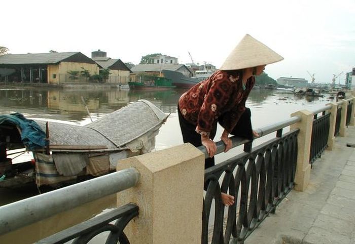 Bà Lan, một người dân xóm đò trèo qua hàng rào mới làm bên bờ sông để lên phố. Phía sau bà, có thể thấy một khúc sông trống trải, phía xa là các cần cẩu của một nhà máy đóng tàu. Ảnh chụp năm 2022