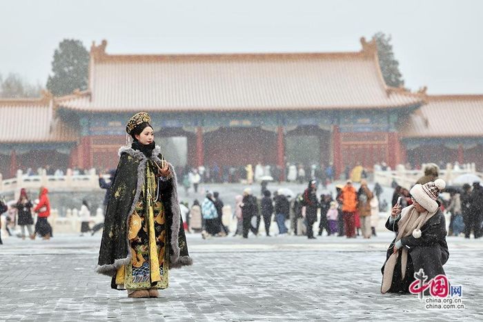  Người dân và du khách nô nức tới Tử Cấm Thành đón đợt tuyết đầu mùa. Nữ du khách diện trang phục hoàng hậu thời xưa, áo choàng dài thêu họa tiết cầu kỳ và mũ đội truyền thống, đứng tạo dáng trên nền quảng trường phủ lớp tuyết mỏng. 