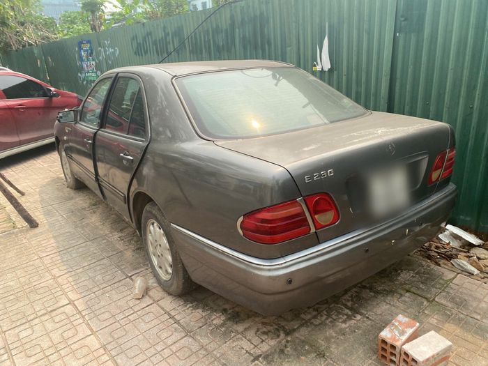Cách đó không xa, chiếc Mercedes-Benz E230 khá hơn khi vẫn giữ được hình dạng ban đầu. Lớp sơn bị bong tróc, gương chiếu hậu hư hỏng và đèn pha đã ố vàng, mờ đục. Toàn bộ xe đóng kín bởi bụi dày nhưng không hỏng hóc nhiều về ngoại quan.