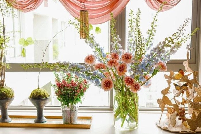 Bên khung cửa sổ ngập nắng, bình hoa thược dược cam san hô kết hợp cùng phi yến xanh lơ tạo nên một bản giao hưởng màu sắc thanh tao. Những chậu cây tiểu cảnh bonsai phía đối diện giúp cân bằng lại không gian bằng nét xanh rì, dịu mắt.
