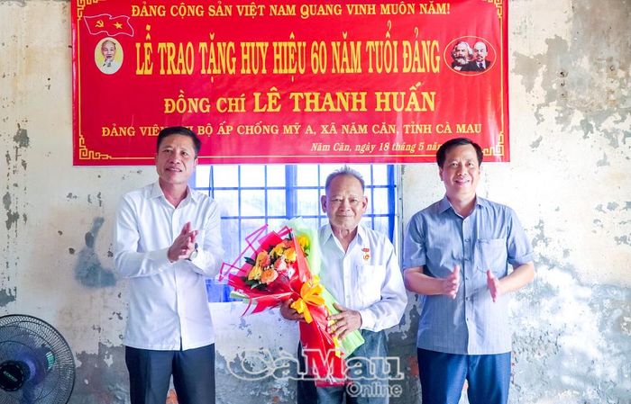 Đồng chí Trần Hữu Phước, Trưởng ban Nội chính Tỉnh ủy và đồng chí Huỳnh Minh Kiên, Bí thư Đảng ủy, Chủ tịch HĐND xã Năm Căn, trao Huy hiệu 60 năm tuổi Đảng và tặng hoa chúc mừng đồng chí Lê Thanh Huấn.