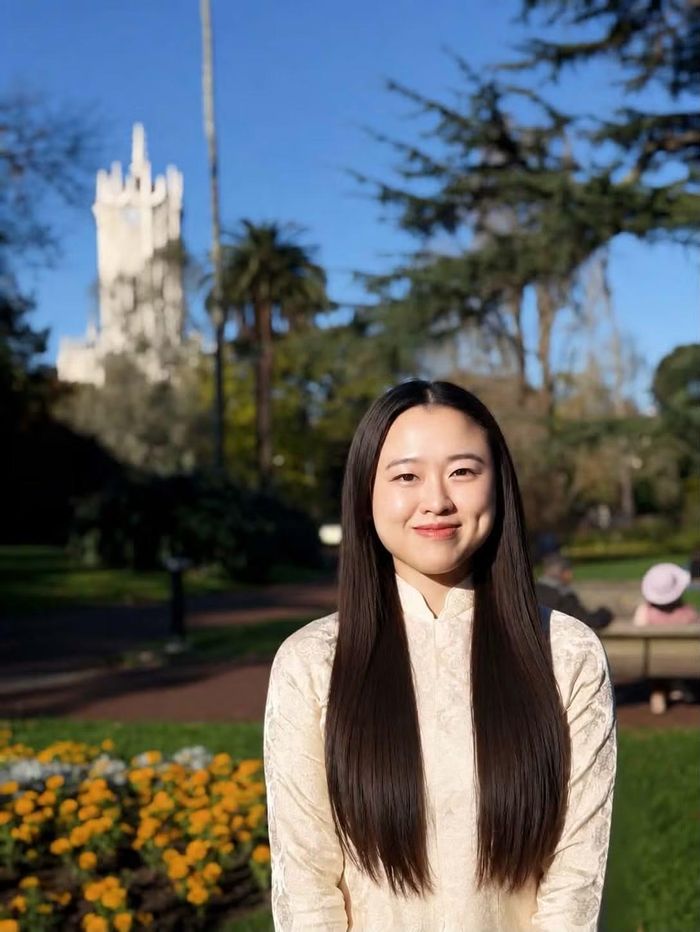 Thùy Nhung trong tà áo dài truyền thống tại khuôn viên Đại học Auckland, phía sau là Clock Tower, công trình biểu tượng mang dấu ấn lịch sử của nhà trường.