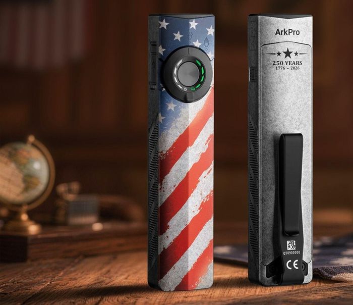 The Olight ArkPro Stars & Stripes Edition flashlight has a thumb switch with a three-way selector dial on the front and a metal pocket clip on the back.