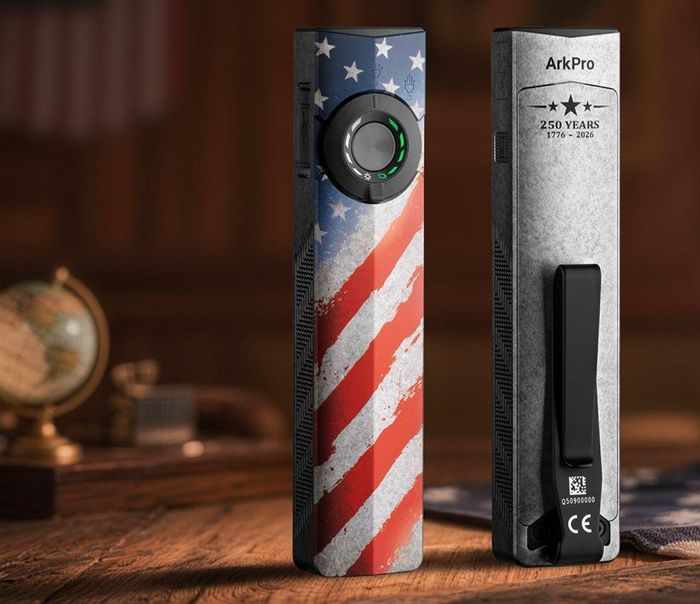 The Olight ArkPro Stars & Stripes Edition flashlight has a thumb switch with a three-way selector dial on the front and a metal pocket clip on the back.