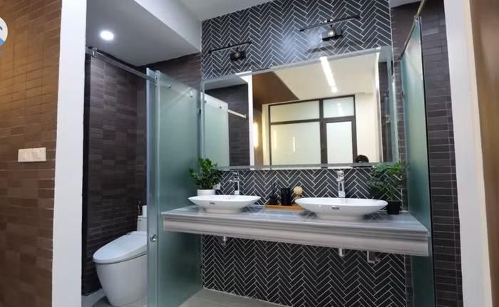 Khu vực rửa tay được bố trí ngay bên ngoài với bồn rửa mặt, gương lớn và tủ lavabo gọn gàng, giúp tối ưu không gian và tăng tính thẩm mỹ. Toàn bộ sàn và tường đều được ốp gạch cao cấp có khả năng chống trơn trượt, chống ẩm và kháng khuẩn, mang lại cảm giác an toàn và sạch sẽ