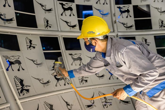 Một nhóm công nhân tỉ mỉ sơn hoàn thiện bề mặt quả cầu màu trắng xám, điểm nhấn của biểu tượng trung tâm. Từng lớp sơn được xử lý kỹ nhằm bảo đảm độ bền và tính thẩm mỹ trước khi công trình hoàn thiện.