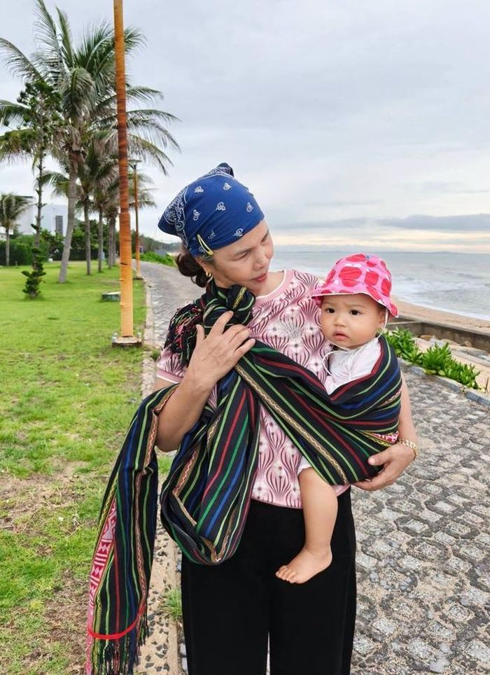 Mẹ H'Hen Niê bế cháu ngoại không rời trong chuyến nghỉ dưỡng của gia đình ở biển Phước Hải. Từ quê Tây Nguyên xuống thành phố, bà mang theo chiếc địu dệt bằng vải thổ cẩm để địu cháu gái Harley suốt hành trình.