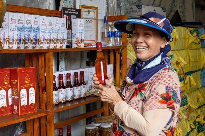 Bà Phan Thị Ngọc Bích, chủ cơ sở sản xuất nước mắm Hương Làng Cổ, giới thiệu mắm ruốc với một người hàng xóm. Theo bà, dù sẵn sàng thử nghiệm sản phẩm mới, nước mắm cá cơm truyền thống vẫn là hương vị đặc trưng của Nam Ô. Ảnh: Xuân Quý - TTXVN