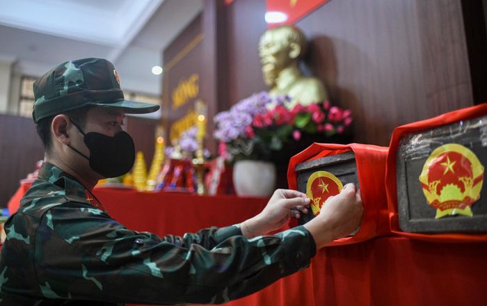  Trong không gian trang nghiêm, một cán bộ Đội Quy tập hài cốt liệt sĩ cẩn thận dán Quốc huy lên tiểu quách chứa hài cốt. Mỗi tiểu quách được bọc vải đỏ, sắp đặt ngay ngắn bên cạnh bàn thờ Chủ tịch Hồ Chí Minh và Quốc kỳ trong Nhà Tưởng niệm. Đây là bước cuối trong quy trình an vị hài cốt trước khi lấy mẫu giám định ADN và lưu giữ theo quy định. 