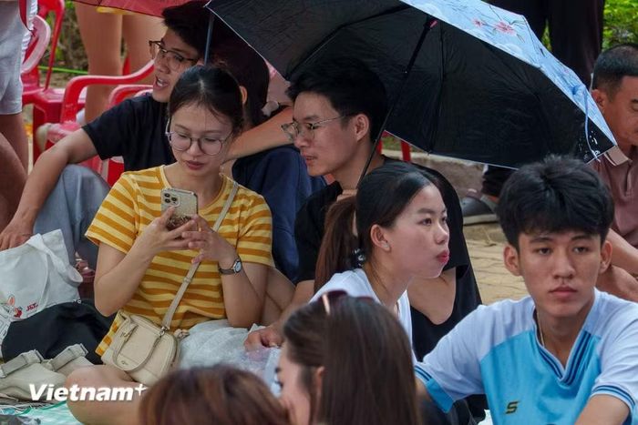 Các bạn trẻ mang theo ô dù, tranh thủ nghỉ ngơi, dùng điện thoại giải trí trong thời gian chờ đợi. (Ảnh: Thanh Phong/Vietnam+)
