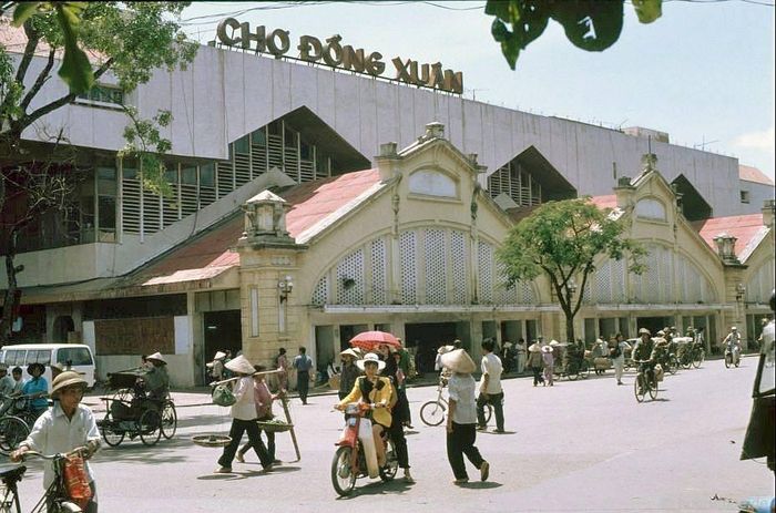  Chợ Đồng Xuân năm 1992 mang đậm nhịp sống Hà Nội thời kỳ mới chuyển mình. Mặt tiền rõ nét kiến trúc Pháp với vòm cong, cửa thông gió hoa văn và mái ngói cũ. Trước chợ đông đúc người dân qua lại, xen lẫn tiểu thương mang dép nhựa, đội nón lá với đôi quang gánh hoặc bao tải hàng. 