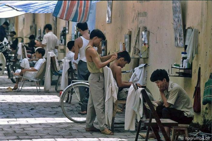  "Phố cắt tóc" trên vỉa hè phố Quang Trung, đoạn giữa phố Bà Triệu và Trần Quốc Toản, vào khoảng năm 1991-1993. Những ô gạch lát vỉa hè, bức tường vàng cũ kỹ cùng các quầy cắt tóc đơn sơ tạo nên khung cảnh bình dị, đặc trưng của Hà Nội thời bấy giờ. Mỗi quầy chỉ có một chiếc gương treo tường, vài chiếc kéo, lược, một ghế gỗ cho khách và đôi khi thêm lọ phấn rôm hoặc keo vuốt tóc tự pha. Khách chủ yếu là nam giới với những kiểu tóc đơn giản. Một lần cắt tóc hoặc cạo râu diễn ra nhanh chóng, chi phí chưa đến một USD. 
