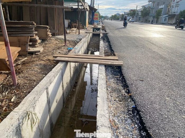 Việc thi công kéo dài cũng khiến cuộc sống của nhiều hộ dân bị đảo lộn. Chị Nga cho biết gia đình sống ngay mặt đường nhưng đến nay vẫn chưa được hoàn trả mặt bằng để làm cổng, lối ra vào nhà còn dang dở, gây nhiều bất tiện trong sinh hoạt hằng ngày. Trong ảnh, người dân phải dùng ván gỗ bắc qua những đoạn mương không nắp để ra vào.