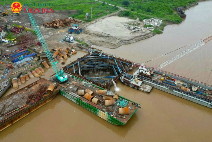 Khu vực thi công nhộn nhịp với hệ thống sà lan, cần cẩu và đường công vụ kéo dài giữa lòng sông