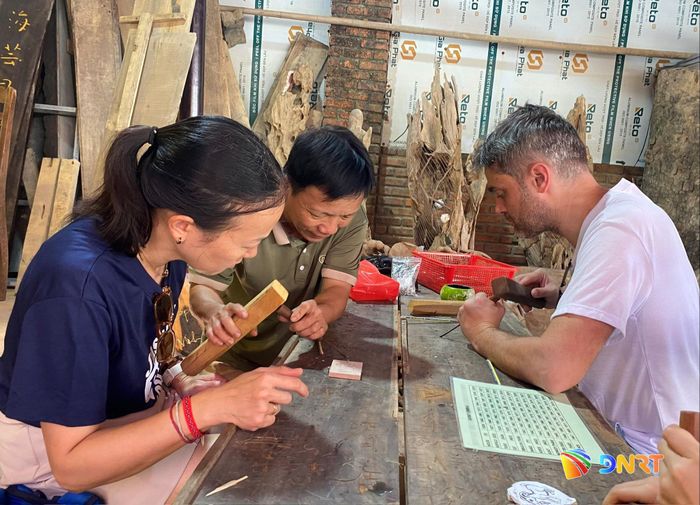 NNND Huỳnh Sướng (giữa) hướng dẫn du khách nước ngoài trải nghiệm chạm trổ nghề mộc Kim Bồng. Ảnh: VĨNH LỘC
