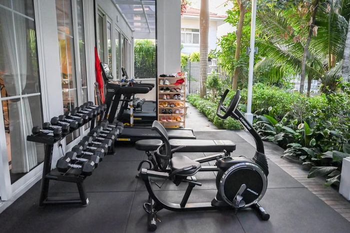  Thay vì bố trí phòng gym kín, gia đình dành một khoảng có mái che bên hông nhà cho các thiết bị tập luyện. Khu vực này có máy chạy bộ, xe đạp, ghế tập và giá tạ. Phía bên ngoài là mảng cây xanh được trồng dọc lối đi. Cách bố trí giúp khu tập luyện có ánh sáng tự nhiên và kết nối trực tiếp với sân vườn. Thu Hương cho biết mỗi sáng cô dành khoảng hai giờ cho việc vận động. Ảnh: NVCC. 