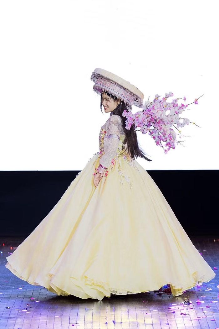 Trên nền váy vàng, những cành hoa đào được thêu và đính kết thủ công, kéo dài từ thân xuống phần váy. Các chi tiết hoa được bố trí theo hướng chuyển động, tạo cảm giác như một nhành đào đang bung nở trên trang phục