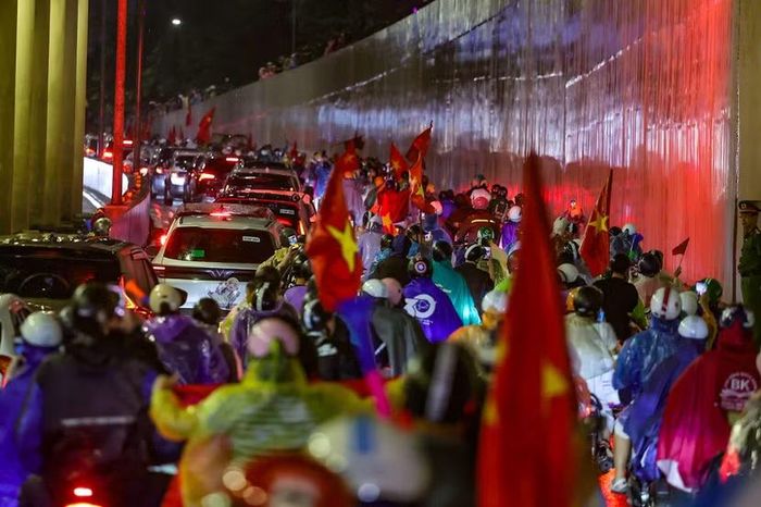 Dòng xe nối đuôi nhau tràn qua hầm chui Kim Liên; ánh đèn hậu và sắc cờ đỏ phản chiếu lên vách hầm bê tông tạo nên khung cảnh rực rỡ đặc trưng của đêm xuống phố ăn mừng.