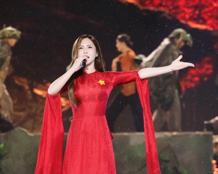 Nữ ca sĩ xuất hiện rực rỡ trong tà áo dài đỏ truyền thống, nổi bật với hình ảnh ngôi sao vàng năm cánh trước ngực—mô phỏng hình ảnh lá Quốc kỳ thiêng liêng.