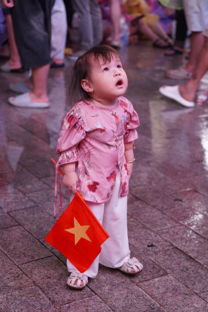 Một em nhỏ chăm chú hướng mắt lên màn hình, trên tay cầm lá cờ Tổ quốc
