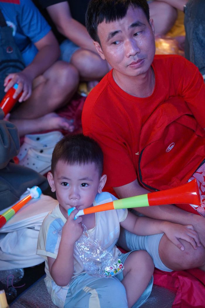 Một cổ động viên nhí cùng người thân ngồi chờ trận đấu, chiếc kèn cổ vũ trên tay