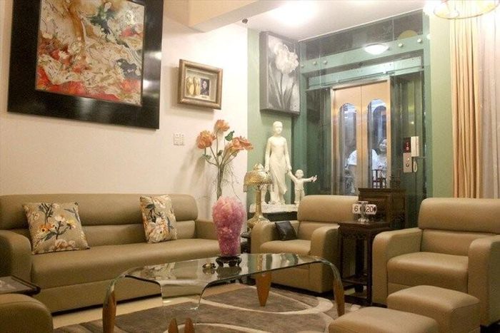 Cận cảnh góc phòng khách với bộ sofa da cao cấp, bàn trà kính và lồng kính thang máy hiện đại phía trong nhà. Sự kết hợp giữa nét cổ điển và tiện nghi hiện đại giúp gia chủ di chuyển dễ dàng giữa các tầng.
