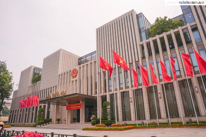 Trước Nhà Quốc hội, những hàng cờ Tổ quốc và cờ Đảng tung bay bên băng rôn chào mừng, cờ phướn và những thảm hoa rực rỡ.