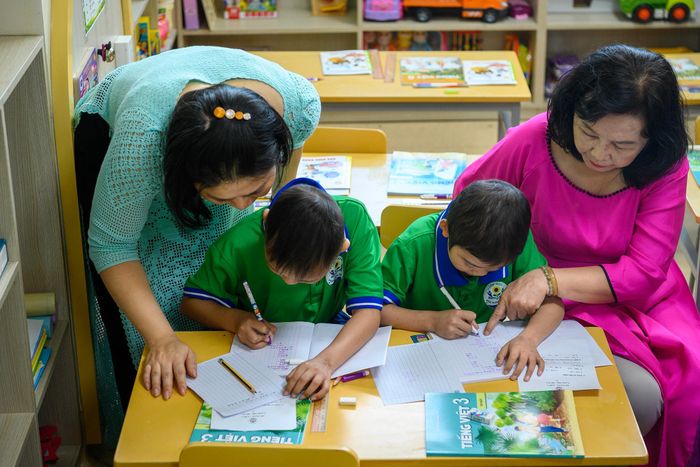 Trong lớp học, cô giáo cầm tay hướng dẫn các em tập viết, kiên nhẫn chỉ bảo từng phép tính.