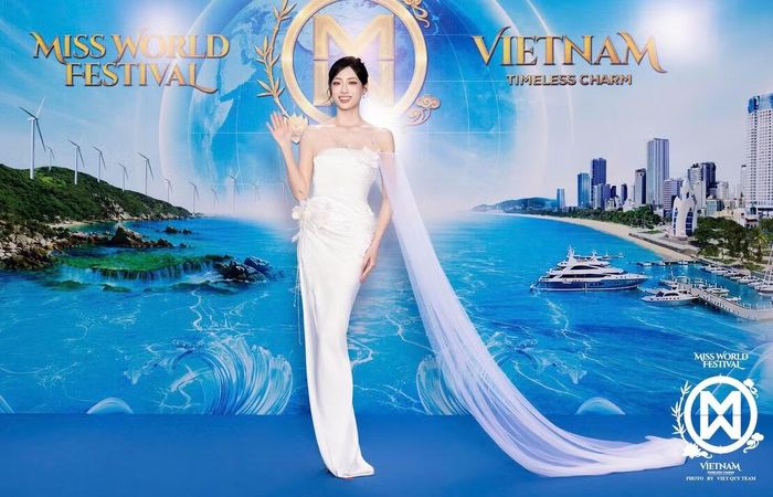 Hoa hậu Lương Thùy Linh diện váy trắng ôm dáng, điểm xuyết chi tiết hoa nổi ở phần thân. Dải voan dài từ vai kéo xuống sàn tạo điểm nhấn. Người đẹp búi tóc cao, kết hợp vòng cổ và hoa tai nhỏ.