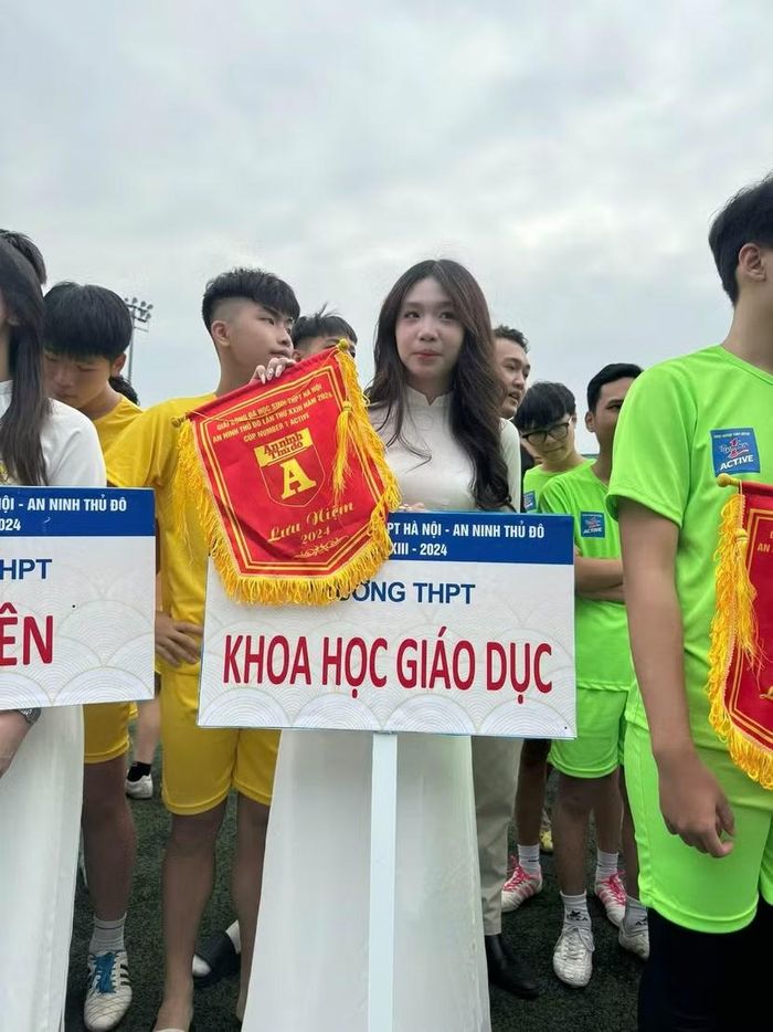 Nguyễn Trần Hà Linh khi còn là cô nữ sinh THPT Khoa học Giáo dục trong tà áo dài thướt tha, cầm biển tên trường tại Lễ khai mạc Giải bóng đá học sinh THPT Hà Nội - An ninh Thủ đô lần thú XXIII năm 2024 Cúp Number 1 Active