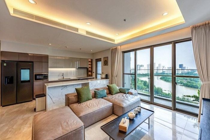 Toàn cảnh phòng khách nhìn ra bờ sông – điểm nhấn đắt giá nhất của căn hộ. Ánh sáng ngập tràn từ hệ cửa kính lớn, mở ra view thành phố và mảng xanh của sông nước phía trước. Bà mẹ một con chia sẻ: "Mẹ chọn một chiếc sofa thật mềm và thật bự để em có thể nhảy nhót lăn lộn tha hồ trên đó".