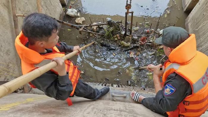 Cảnh sát cơ động bám mép kè bê tông trơn dốc, dùng sào tre phá các mảng rác nén chặt trước vỉ sắt chắn nước.