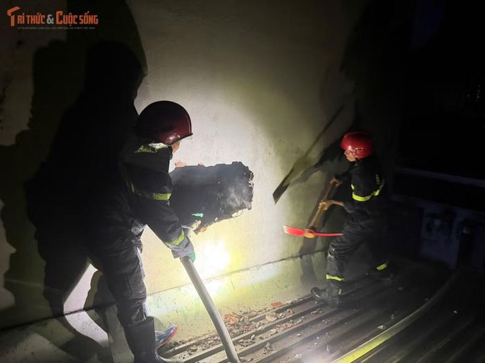 Lính cứu hỏa dùng búa đập tường tạo khoảng trống thoát khói, đồng thời kéo vòi rồng phun nước từ nhiều hướng để dập lửa.
