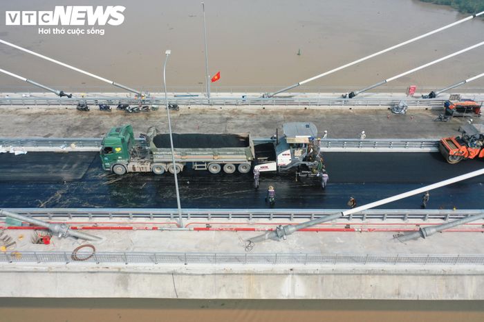 Trên mặt cầu, từng tốp công nhân bám sát dây chuyền. Máy thảm nhựa di chuyển chậm trên từng mét mặt đường, phía sau là công nhân liên tục căn chỉnh, san gạt và kiểm tra độ phẳng của lớp vật liệu.